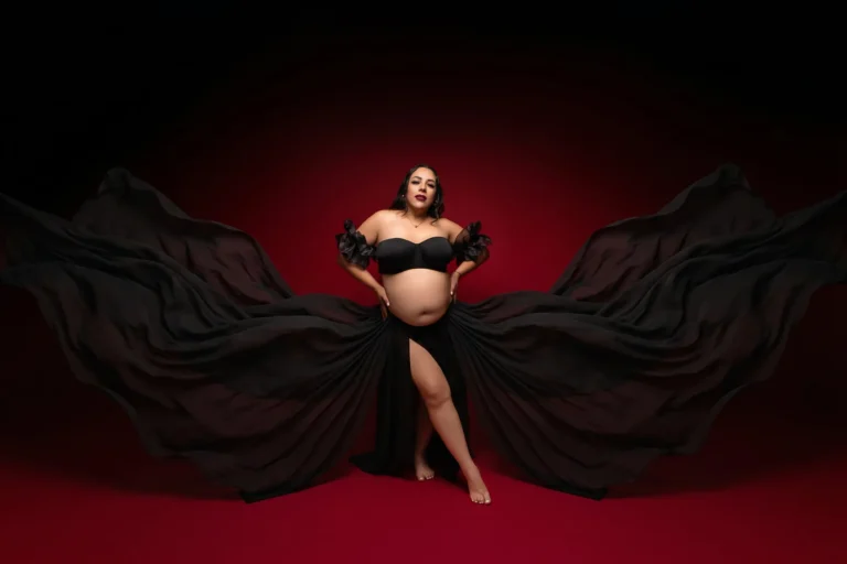 Mujer embarazada con vestido negro y tela en movimiento sobre fondo rojo, sesión de maternidad en estudio en Tijuana.